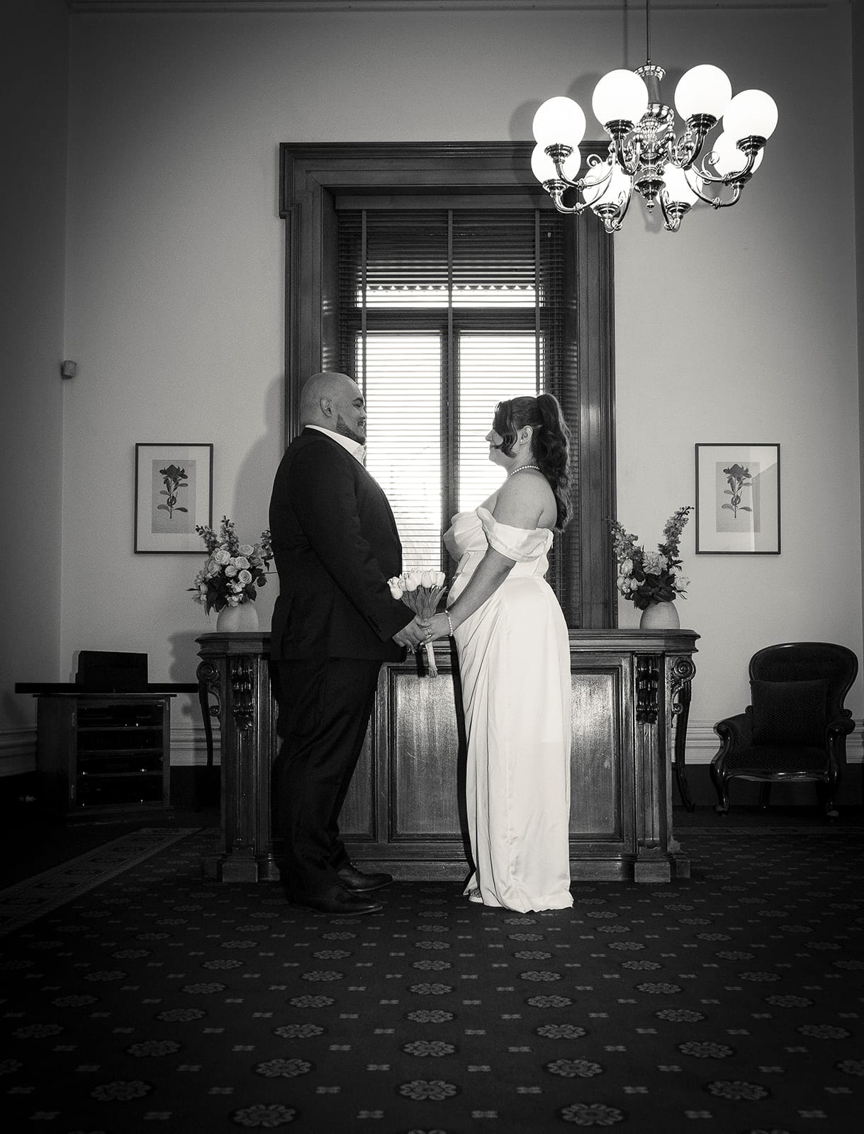 Black and white photo of Martina and Diego holding hands at the registry desk under the chandelier