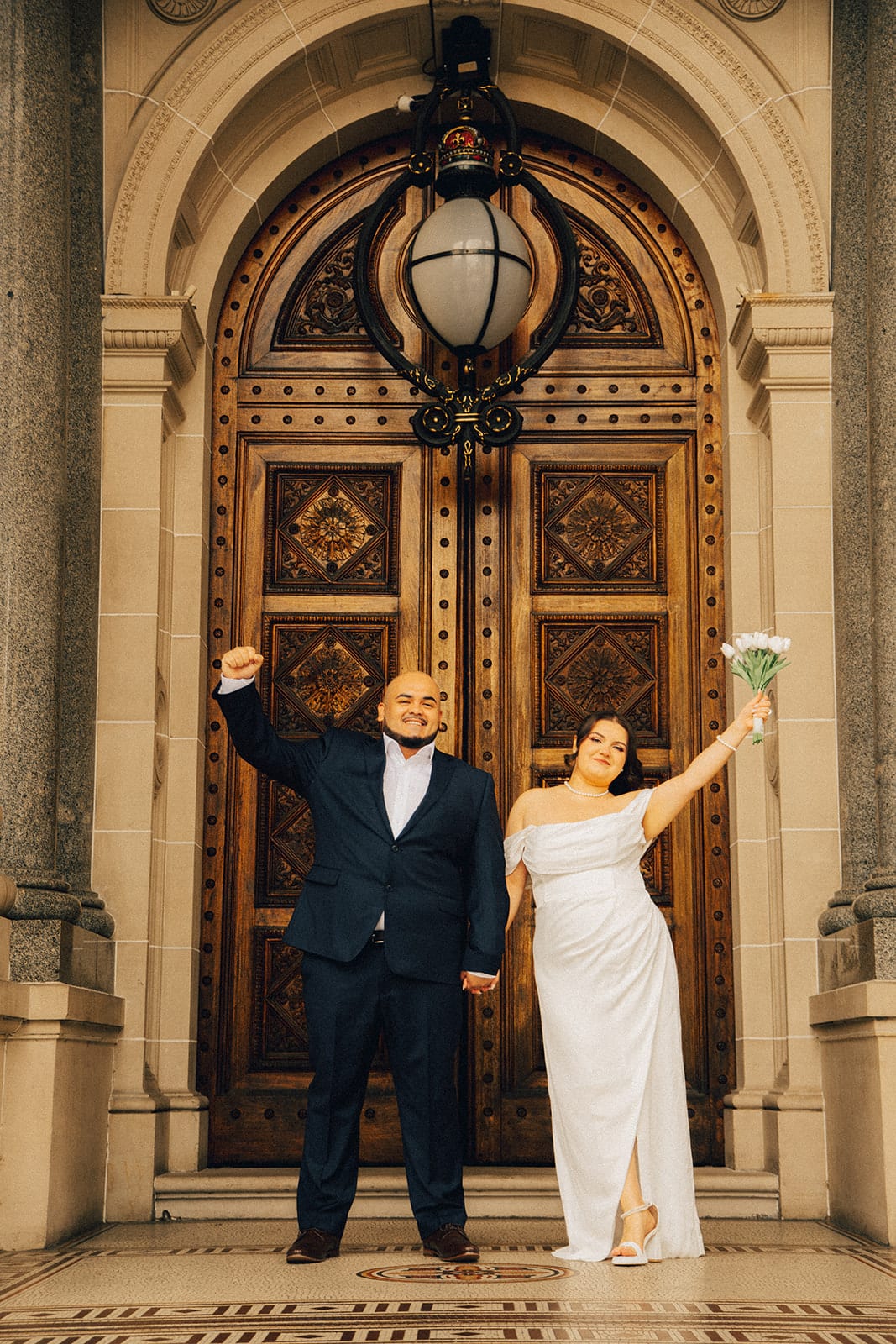 Martina and Diego cheering with their arms in the air in front of the carved doors of Parliament House