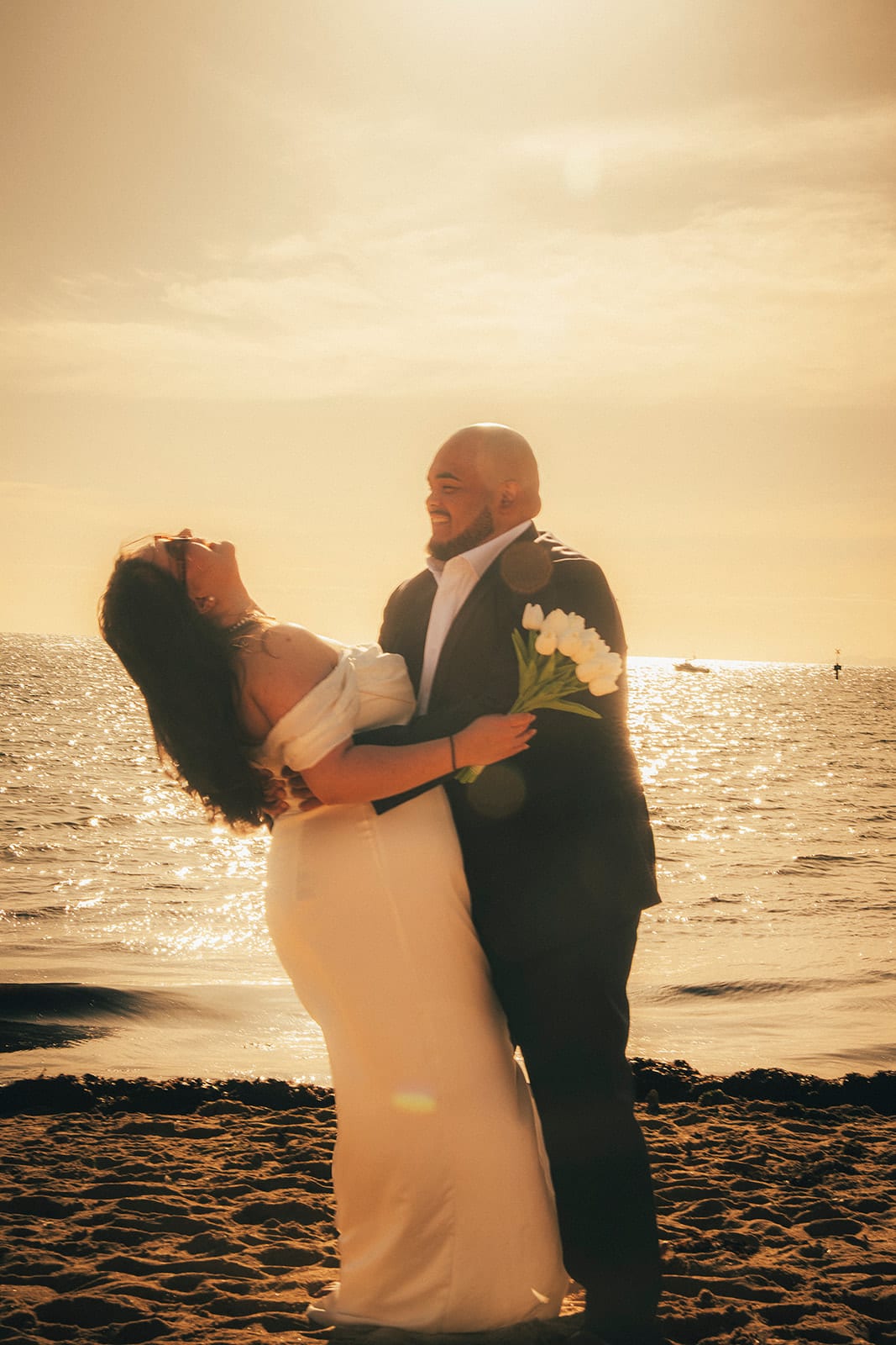 Martina laughing with her head thrown back in Diego’s arms, the low sun over the water behind them
