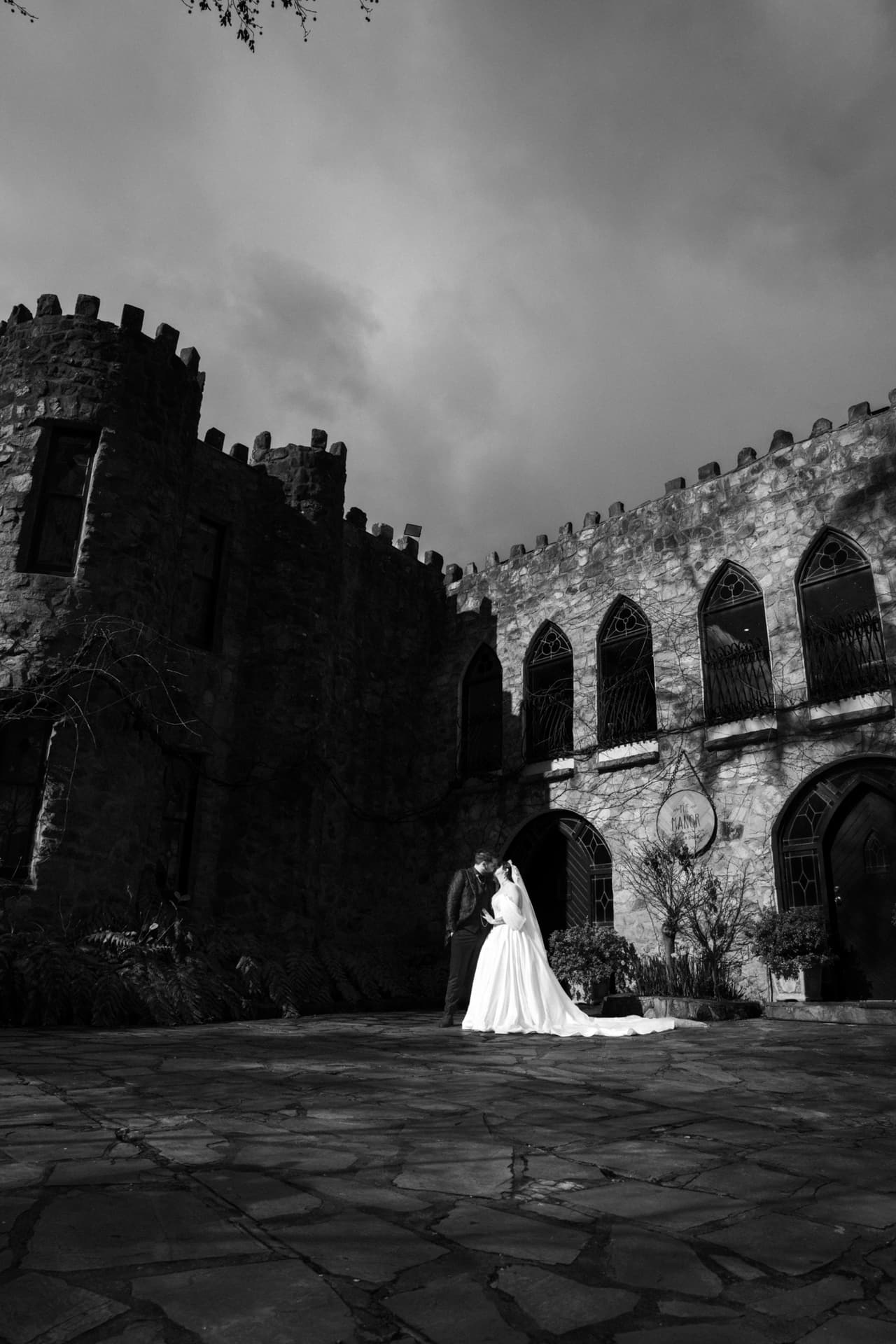 Black and white photo of Porscha and Jordan in the stone courtyard of The Manor, its battlements against a stormy sky