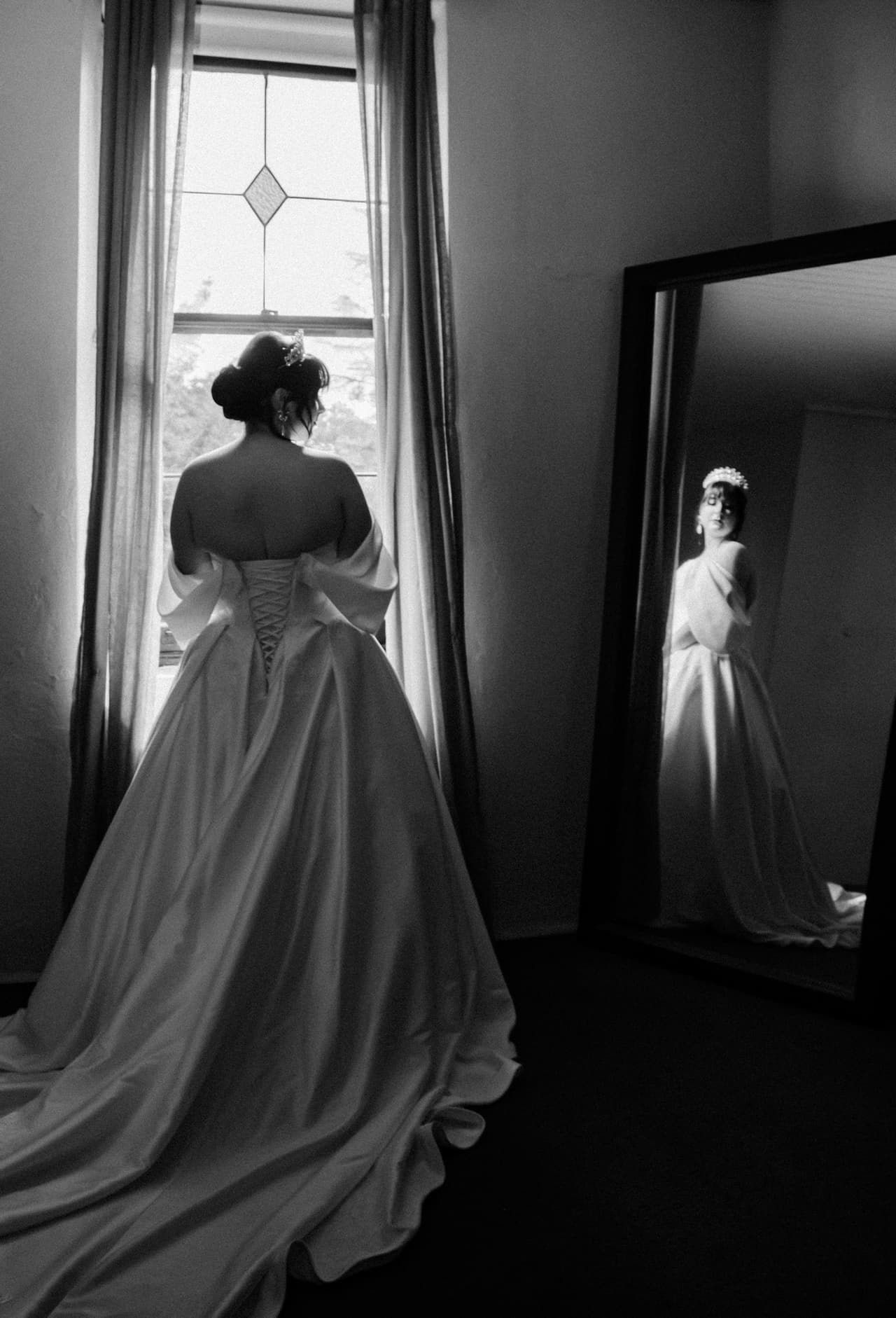 Black and white photo of Porscha at a tall window, her gown’s laced back to the camera and her reflection in a mirror
