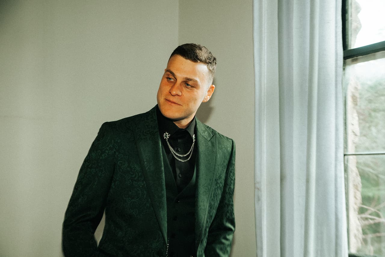 One of the men in a dark green suit, smiling by the window as they get ready