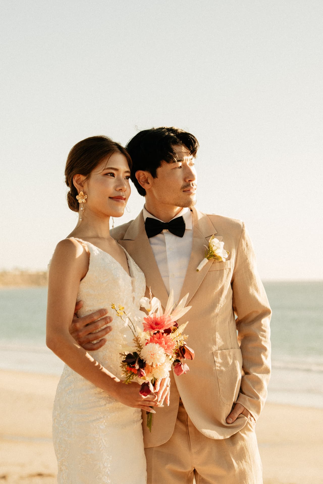 Sayuri and Shuto in a beige suit on the beach in golden light, her bouquet in hand