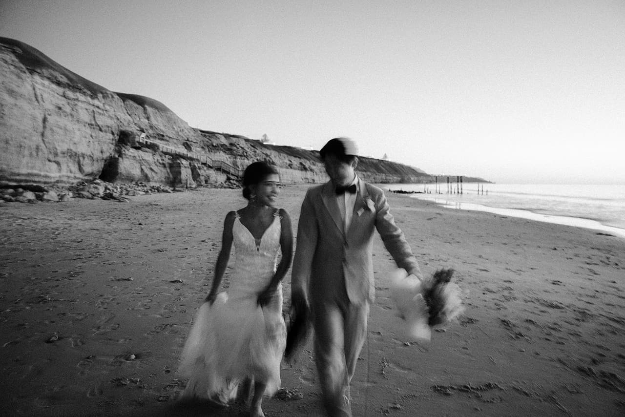 Black and white photo of Sayuri and Shuto walking along the beach at dusk, the cliffs and the jetty posts behind them