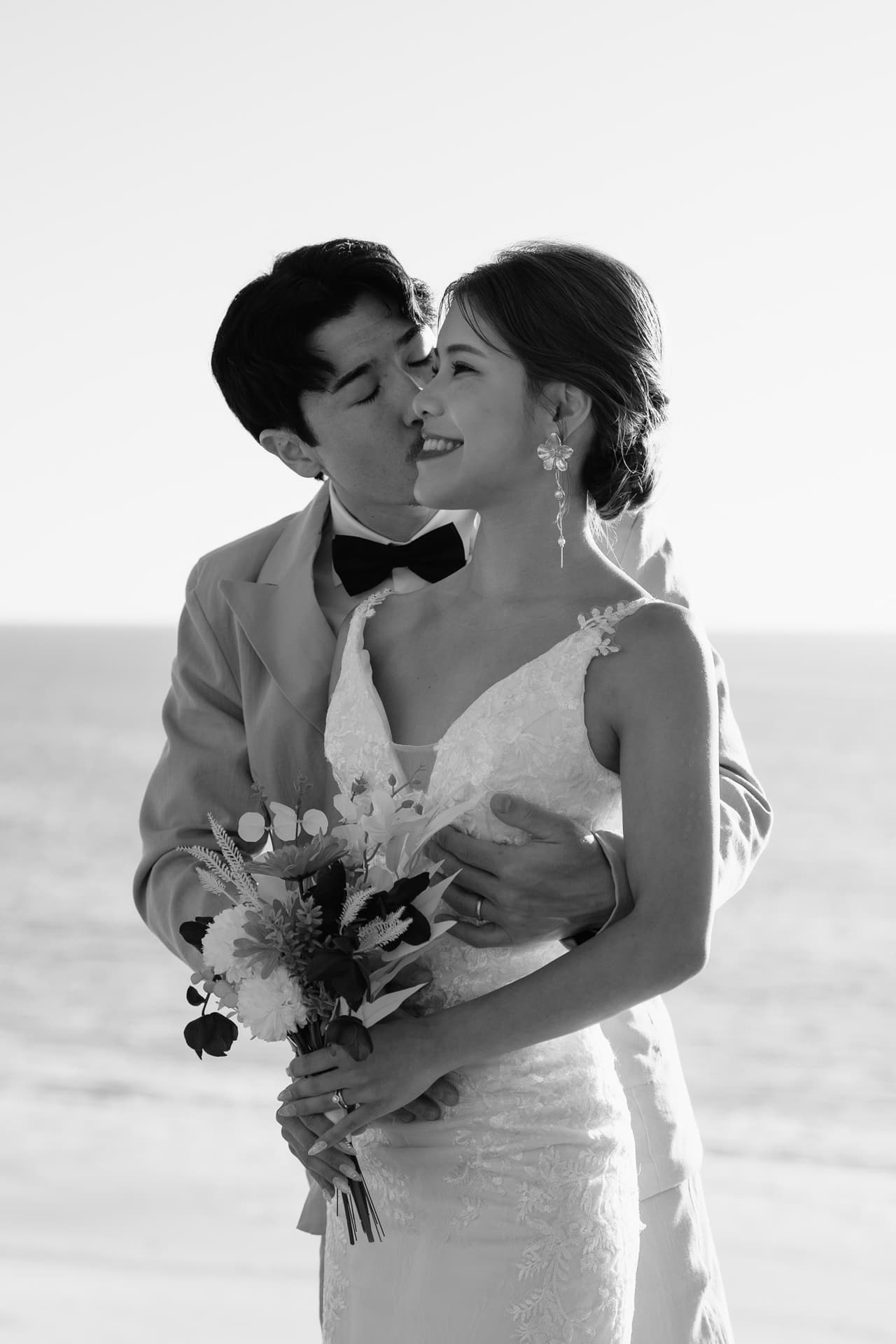 Black and white photo of Shuto kissing Sayuri’s cheek by the sea, her bouquet in hand