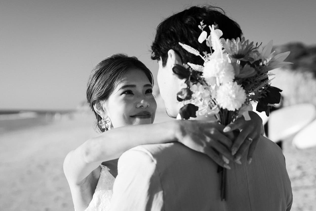 Black and white photo of Sayuri smiling over Shuto’s shoulder as she hugs him, her bouquet behind his head