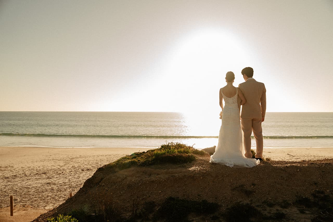 Sayuri and Shuto seen from behind on the sandy point, looking out at the sun over the sea