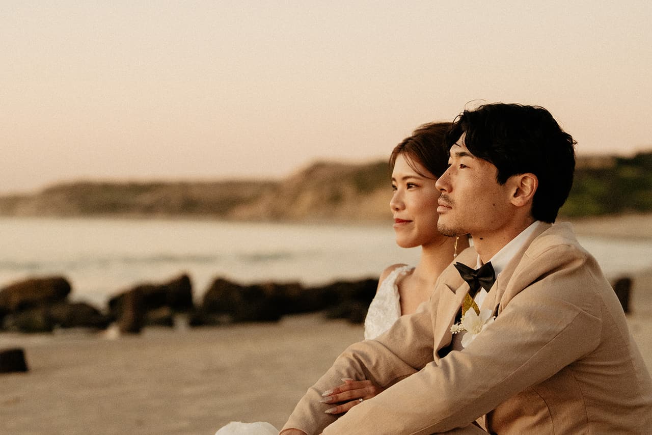 Sayuri and Shuto sitting together on the sand, looking out over the rocks and the sea in the last light