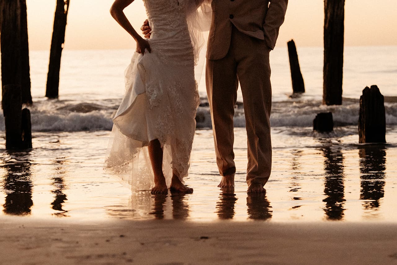 Sayuri holding up her gown as she and Shuto stand barefoot in the shallows among the jetty posts