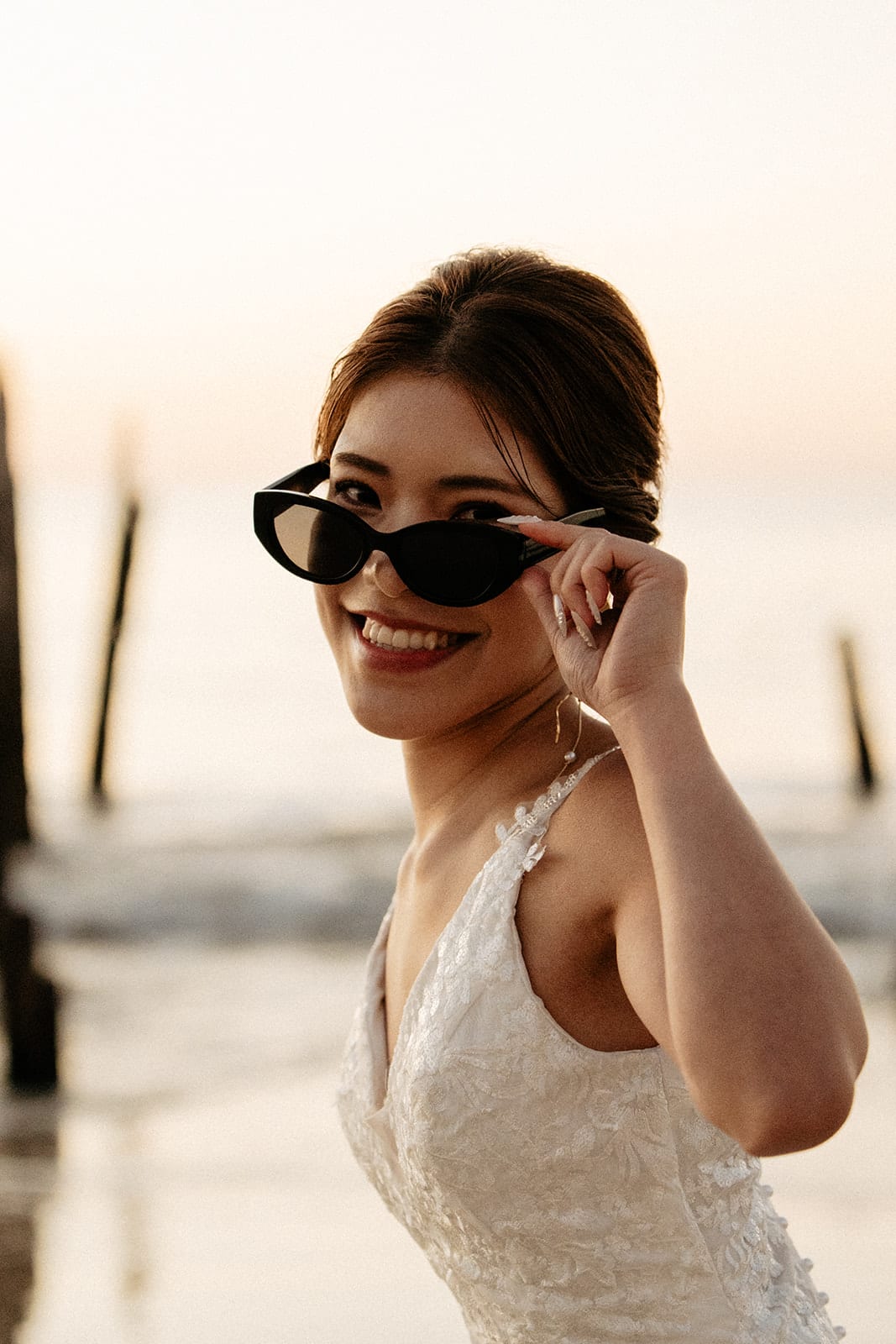 Sayuri smiling as she lowers her sunglasses beside the jetty posts at dusk