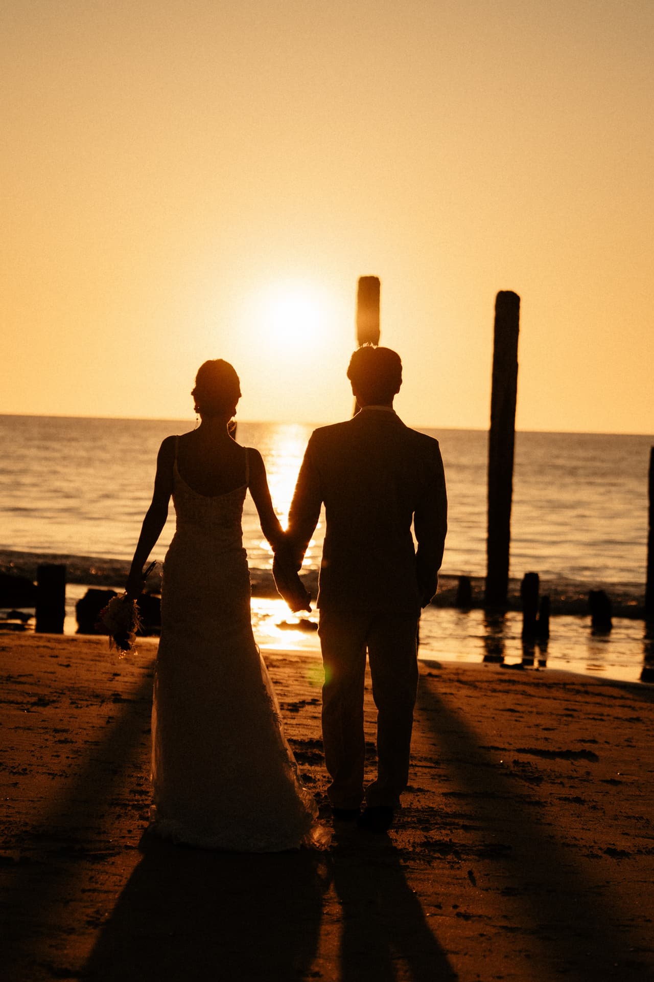 Sayuri and Shuto in silhouette walking hand in hand towards the setting sun beside the old jetty posts