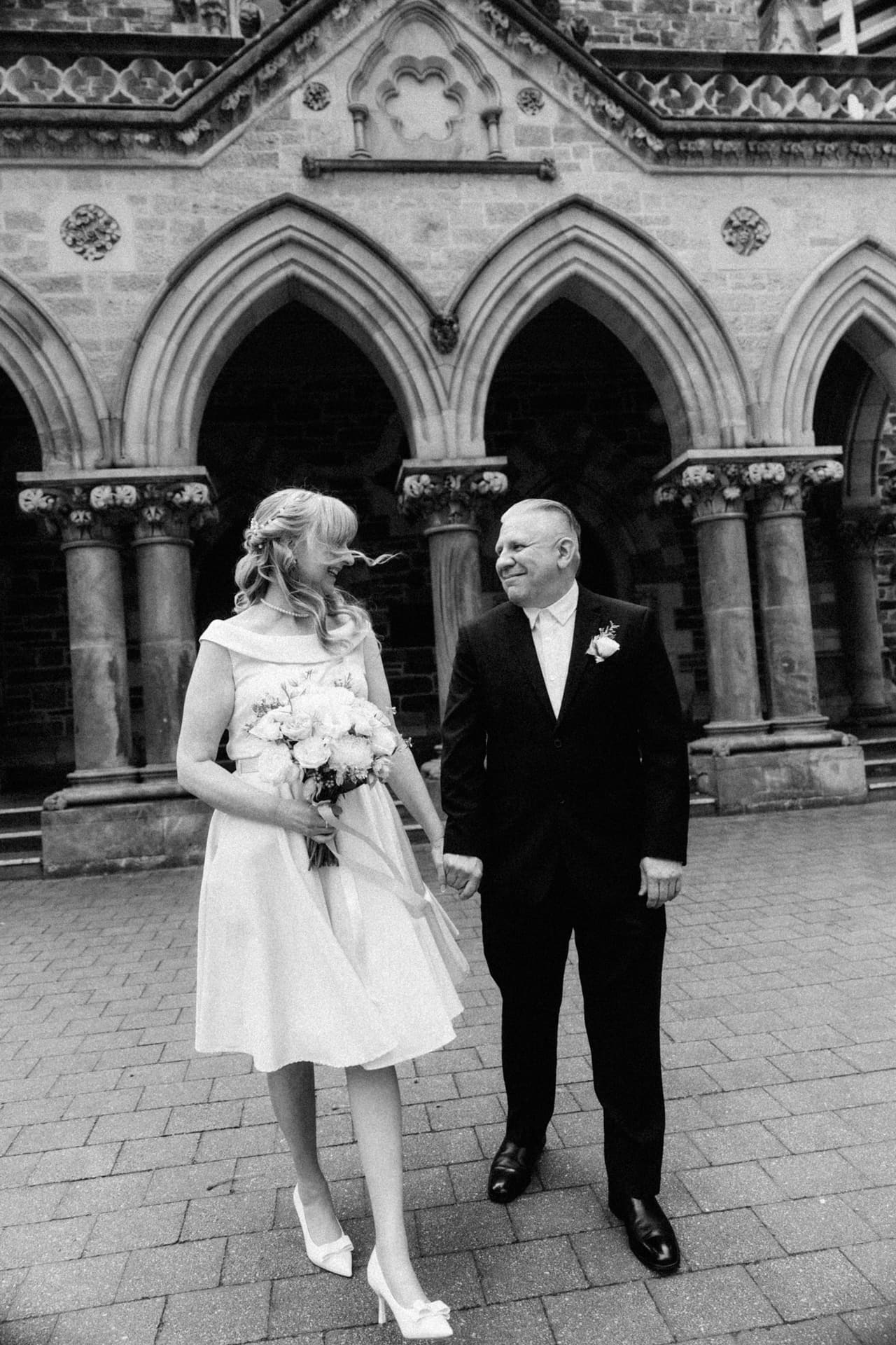 Black and white photo of Terrie and Jon holding hands in front of sandstone Gothic arches, the wind in her hair