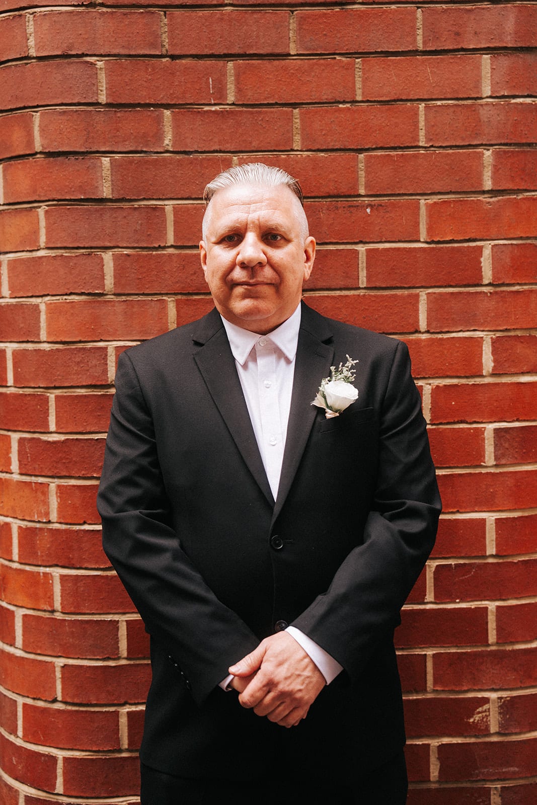 Jon in his black suit with a white rose buttonhole, standing against a red brick wall