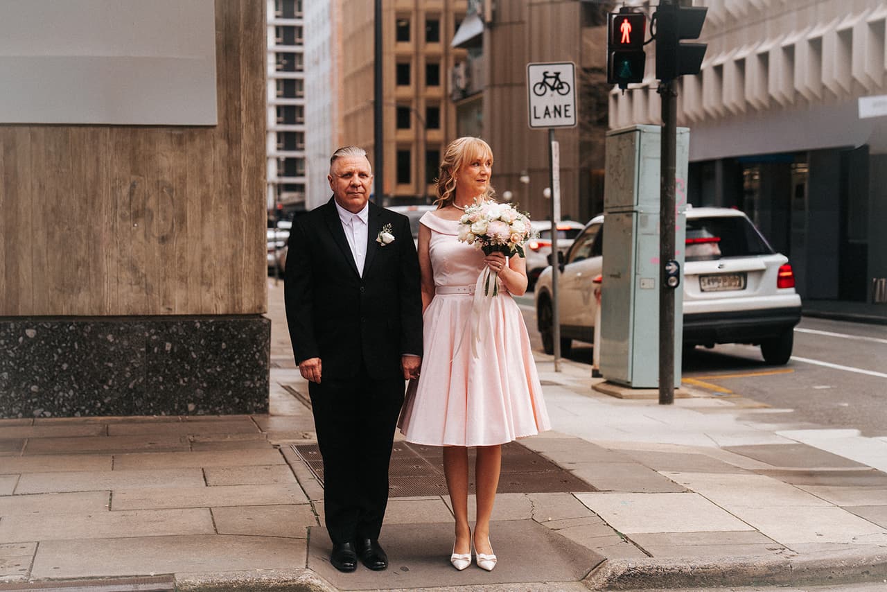 Terrie and Jon standing at the kerb beside a set of traffic lights on a city street