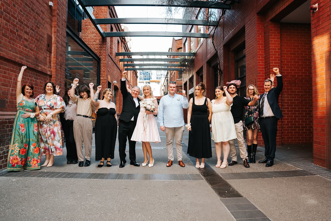 Terrie, Jon and their family and friends cheering with their arms in the air in the laneway outside the registry