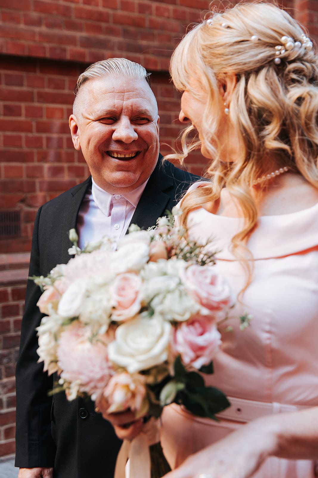 Jon laughing with Terrie against a red brick wall, her bouquet held between them