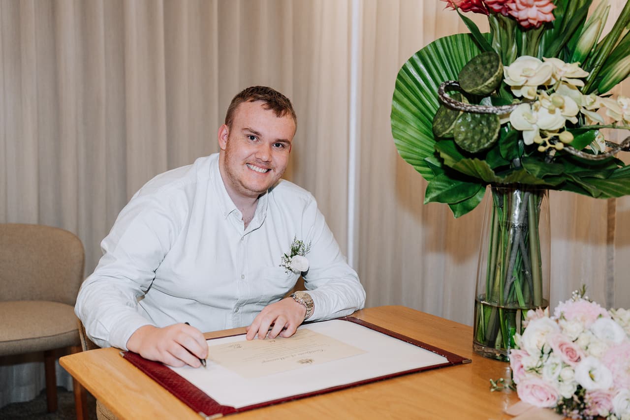 A young man in a pale blue shirt smiling as he signs as a witness at the registry table